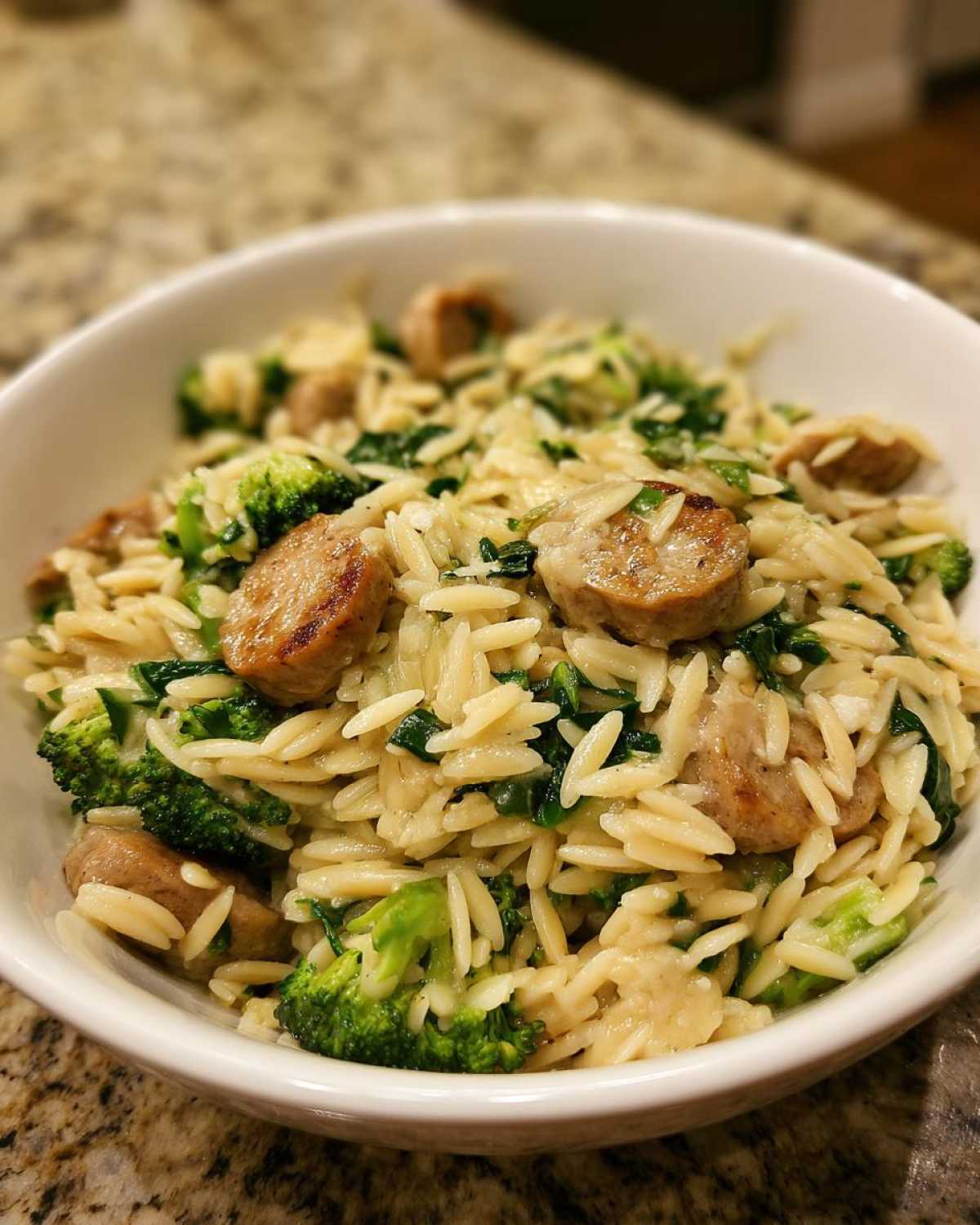 Close-up of a white bowl filled with chicken sausage and broccoli orzo, featuring sliced browned sausage pieces.