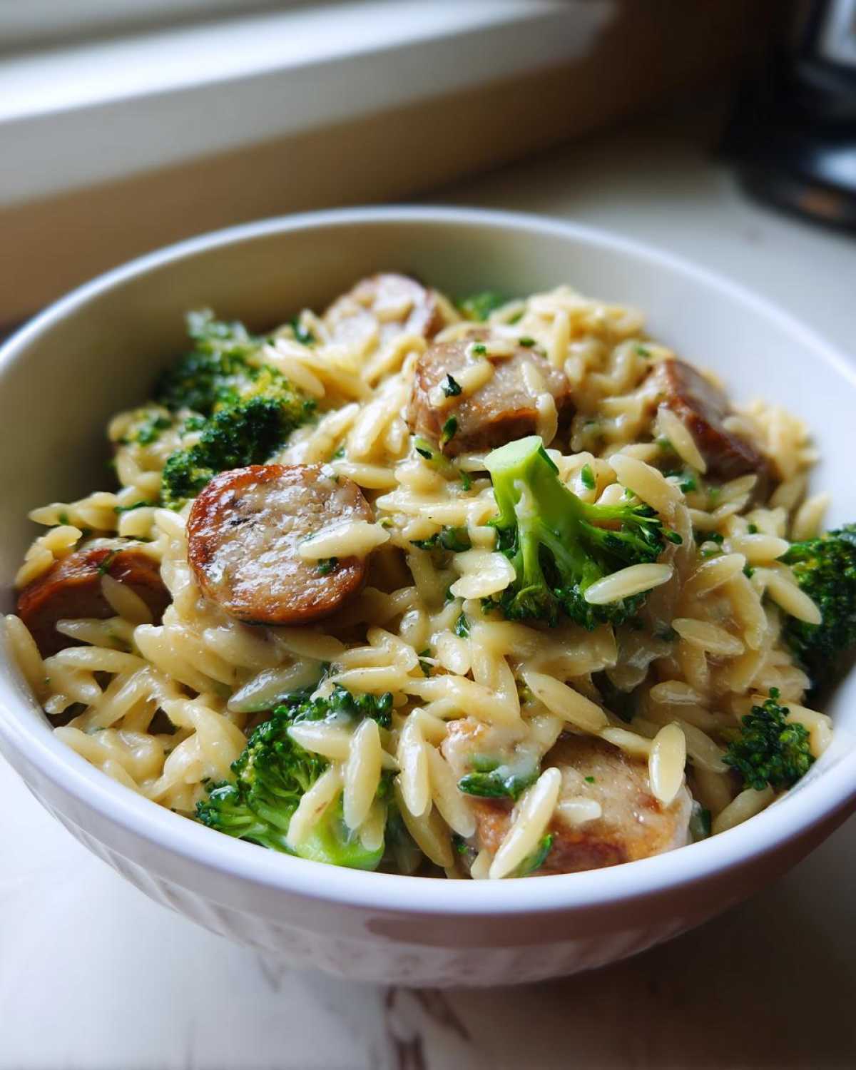 A close-up of a creamy bowl of chicken sausage and broccoli orzo, garnished with fresh herbs.