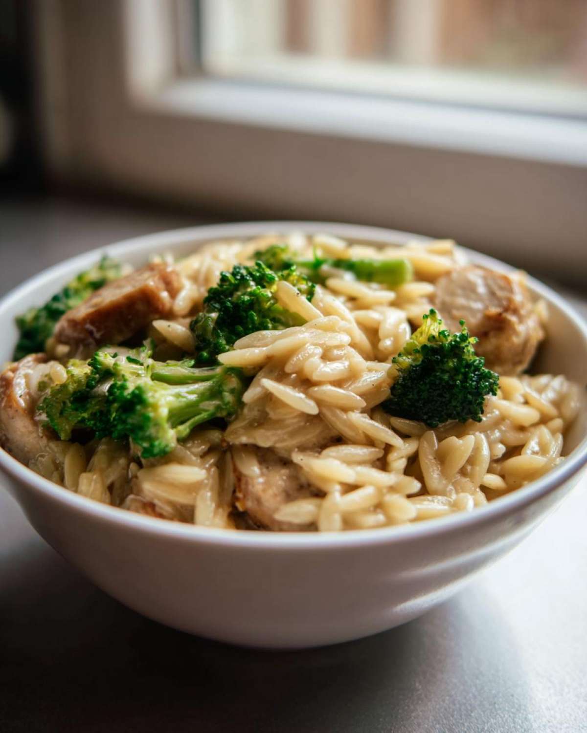 Close-up of creamy chicken sausage and broccoli orzo served in a white bowl near a window.