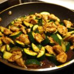 Close-up of chicken zucchini stir fry cooking in a dark skillet with a glossy brown sauce.