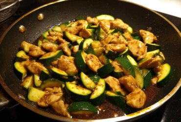 Close-up of chicken zucchini stir fry cooking in a dark skillet with a glossy brown sauce.