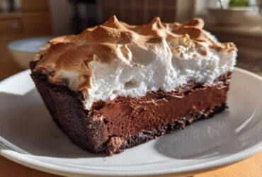 A close-up slice of chocolate meringue pie showing a dark chocolate filling and a tall, toasted meringue topping.