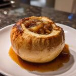 A single, perfectly cooked baked apple filled with spiced, caramelized chunks, resting in its own syrup on a white plate.