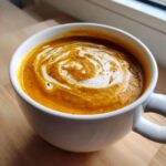 Close-up of creamy butternut squash soup in a white mug, swirled with cream and sprinkled with pepper.
