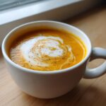 A close-up of creamy, orange butternut squash soup topped with a swirl of cream and cracked black pepper.
