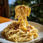 A fork lifting creamy fettuccine noodles from a bowl of caramelized onion pasta, topped with herbs and crispy bits.