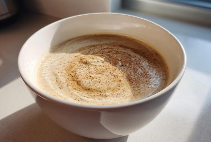 A close-up of creamy cauliflower soup in a white bowl, swirled and dusted with nutmeg or pepper.