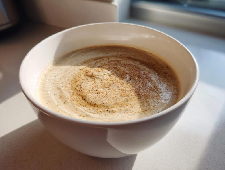 A close-up of creamy cauliflower soup in a white bowl, swirled and dusted with nutmeg or pepper.