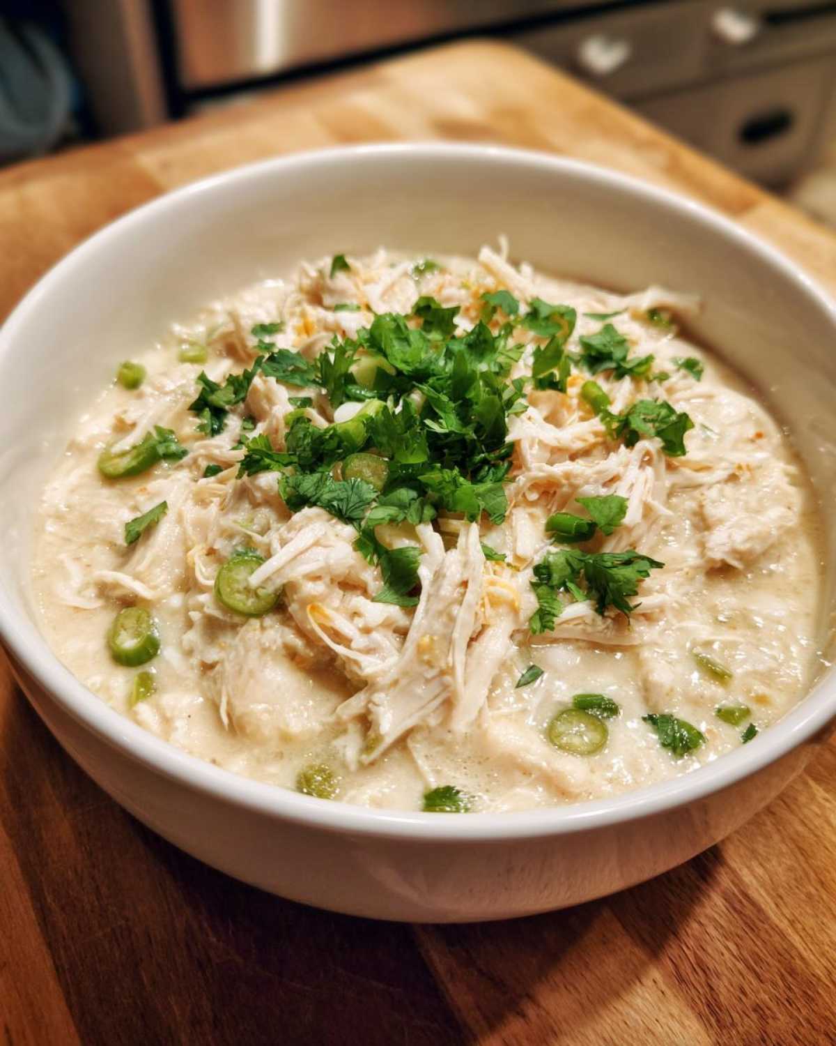 A close-up of a white bowl filled with creamy chicken chili, topped with shredded chicken, cilantro, and green onions.