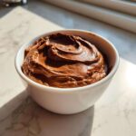 A close-up of rich, creamy chocolate dip swirled in a small white bowl, catching sunlight.