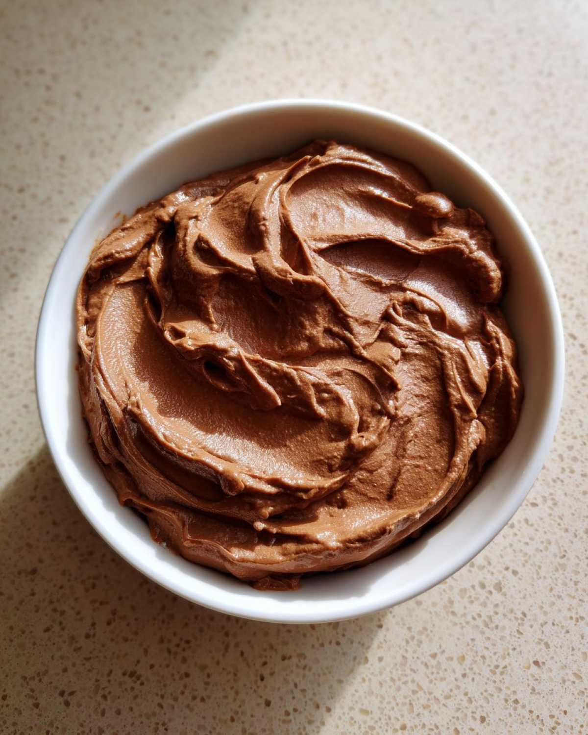 Overhead view of a white bowl filled with thick, creamy chocolate dip, showing swirls on the surface.