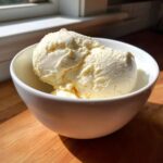 Two scoops of creamy, pale yellow cottage cheese ice cream served in a white bowl on a wooden surface.