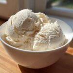 Three scoops of creamy, vanilla-colored cottage cheese ice cream served in a white bowl on a wooden surface.