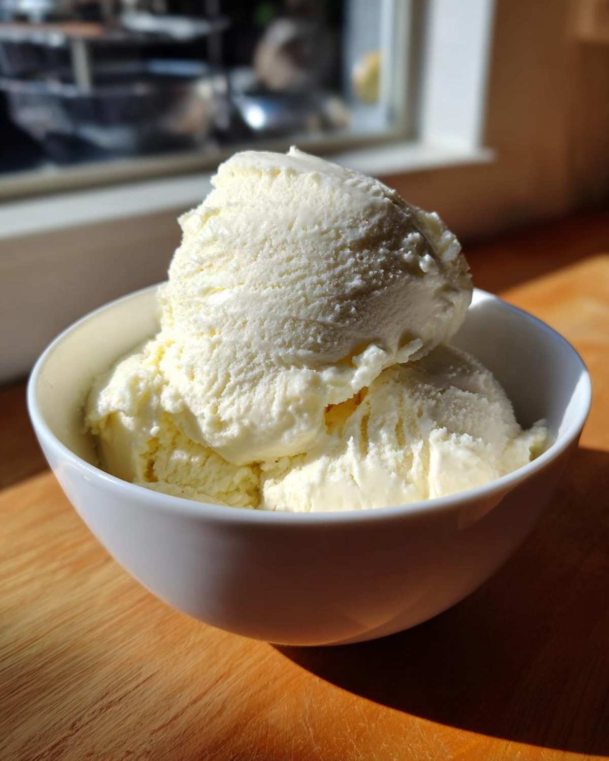 Two scoops of creamy, pale yellow cottage cheese ice cream served in a white bowl on a wooden surface.