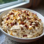A white bowl filled with creamy Neiman Marcus dip, topped generously with shredded cheese, bacon bits, and sliced almonds.