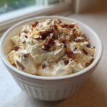 A white bowl filled with creamy Neiman Marcus dip, topped generously with crumbled bacon, shredded cheese, and chopped nuts.