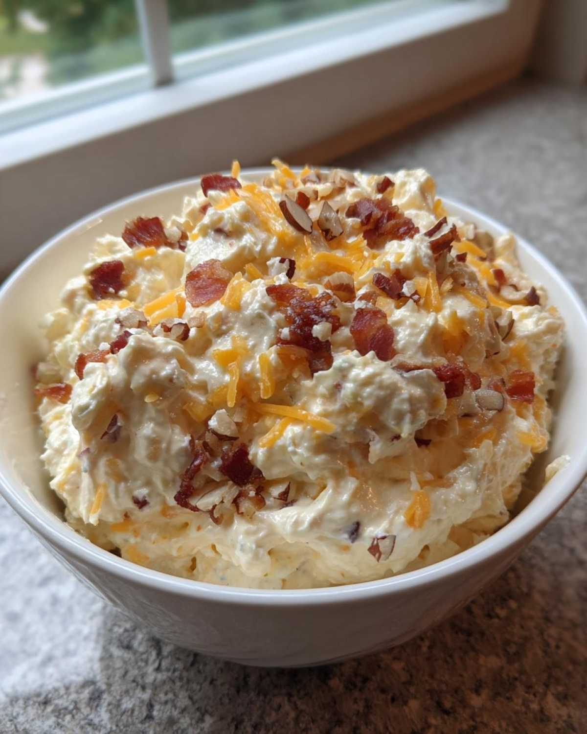 A white bowl filled with creamy Neiman Marcus dip, topped generously with shredded cheddar cheese, bacon bits, and chopped pecans.