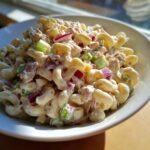 A close-up, bright photo of creamy Tuna pasta salad featuring elbow macaroni, chunks of tuna, red onion, and celery in a white bowl.