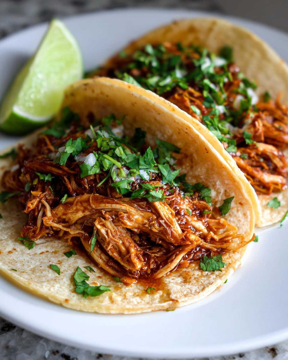 Two soft corn tortillas filled with saucy shredded chicken, topped with cilantro and onion, served with a lime wedge for fresh chicken tacos.