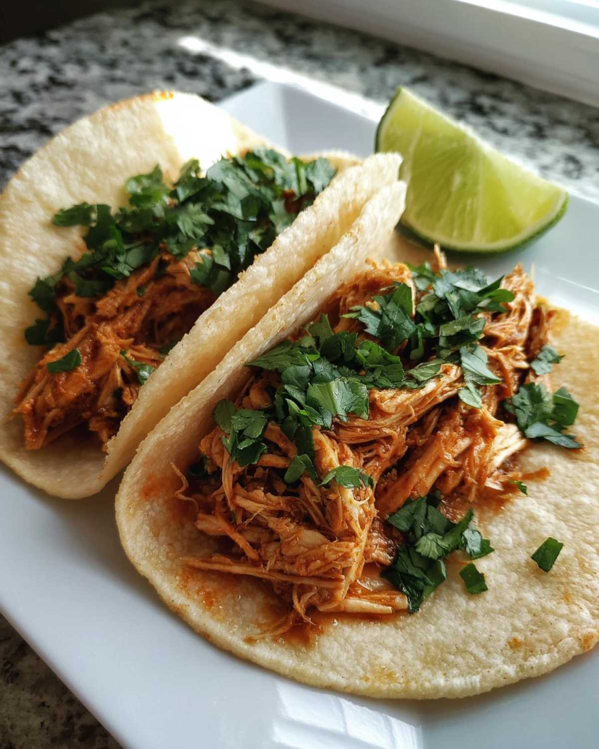 Two soft corn tortillas filled with shredded, saucy chicken and topped generously with fresh cilantro, served with a lime wedge.