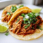 Close-up of two soft corn tortillas filled with saucy shredded chicken tacos, topped with fresh cilantro and onion.