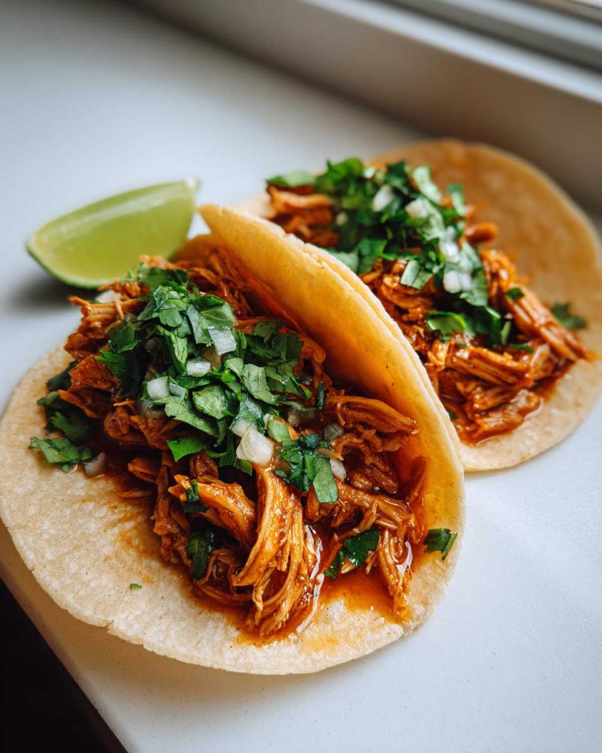 Two soft corn tortillas filled with saucy shredded chicken tacos, topped with fresh cilantro and onion, next to a lime wedge.