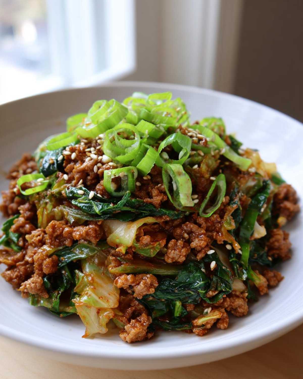 A close-up of a serving of egg roll in a bowl, featuring ground meat, cabbage, and spinach, topped with green onions and sesame seeds.