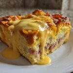 A close-up of a slice of baked eggs benedict casserole topped with rich, dripping hollandaise sauce.