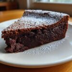A decadent slice of flourless chocolate torte, dusted lightly with powdered sugar, served on a white plate.