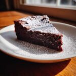 A decadent slice of flourless chocolate torte dusted heavily with powdered sugar, served on a white plate.