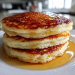 A close-up of three fluffy pancakes stacked high and generously drizzled with golden syrup.