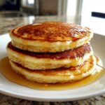 A close-up of three thick, fluffy pancakes stacked high and generously drizzled with golden syrup.