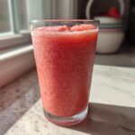 A tall glass filled with a bright red, icy strawberry daiquiri sitting on a sunlit countertop.