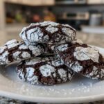 A stack of four fudgy chocolate crinkle cookies heavily dusted with powdered sugar, resting on a white plate.
