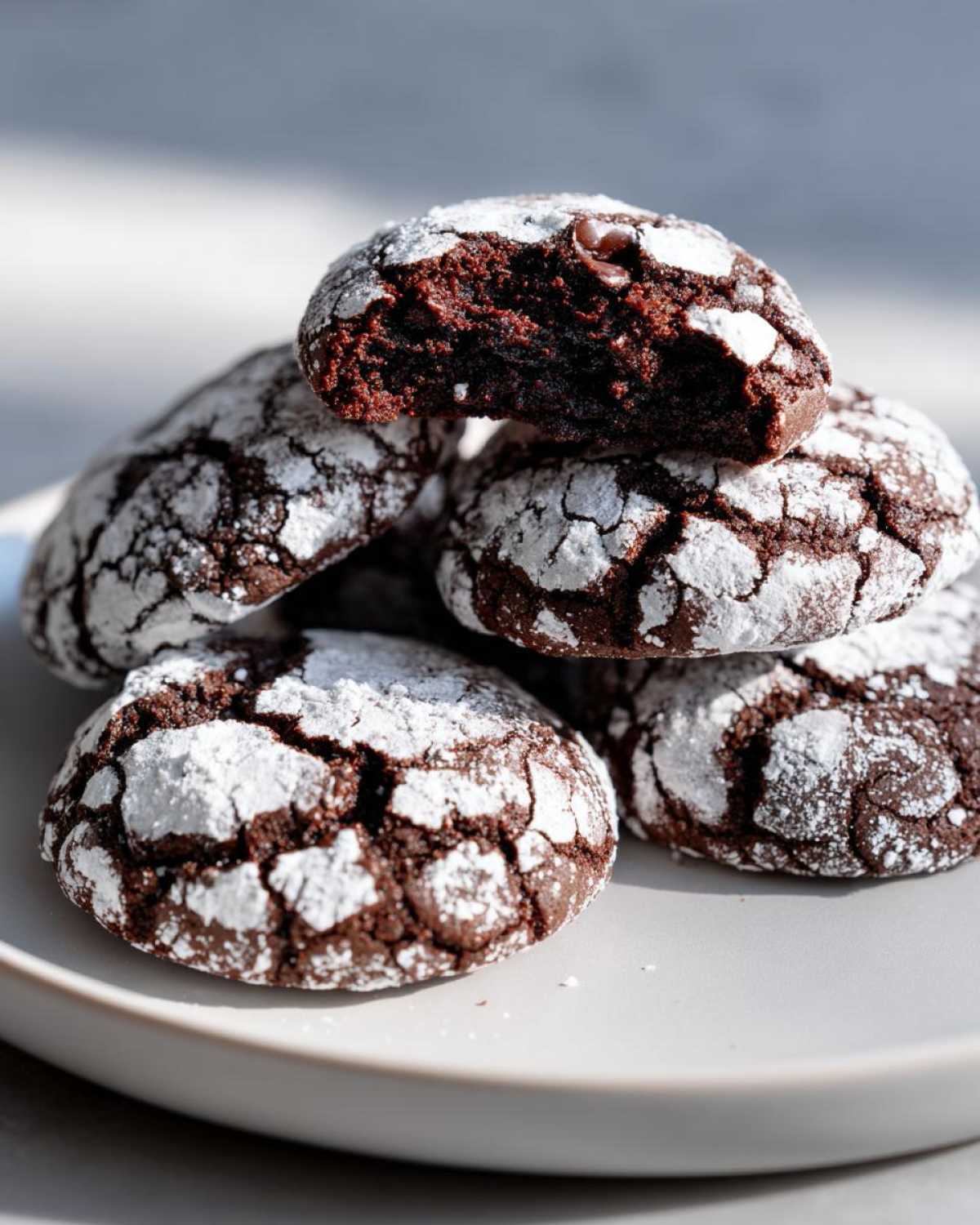 A stack of fudgy crinkle cookies dusted with powdered sugar, with the top cookie broken open to show the rich, dark interior.