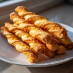 A stack of freshly baked, golden, twisted cheese straws recipe items piled on a white plate.