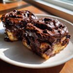 Two decadent chocolate cinnamon rolls heavily topped with glossy, melted chocolate frosting.