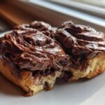 Close-up of two gooey chocolate cinnamon rolls generously topped with thick chocolate frosting on a white plate.