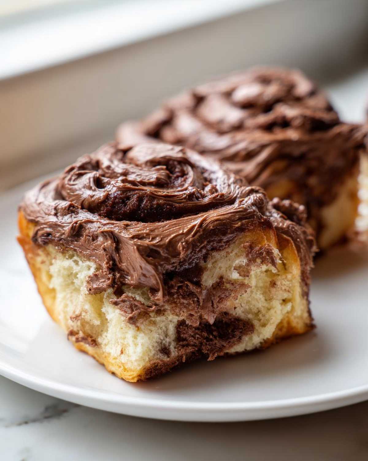 Close-up of a soft, fluffy chocolate cinnamon roll topped with thick, rich chocolate frosting.