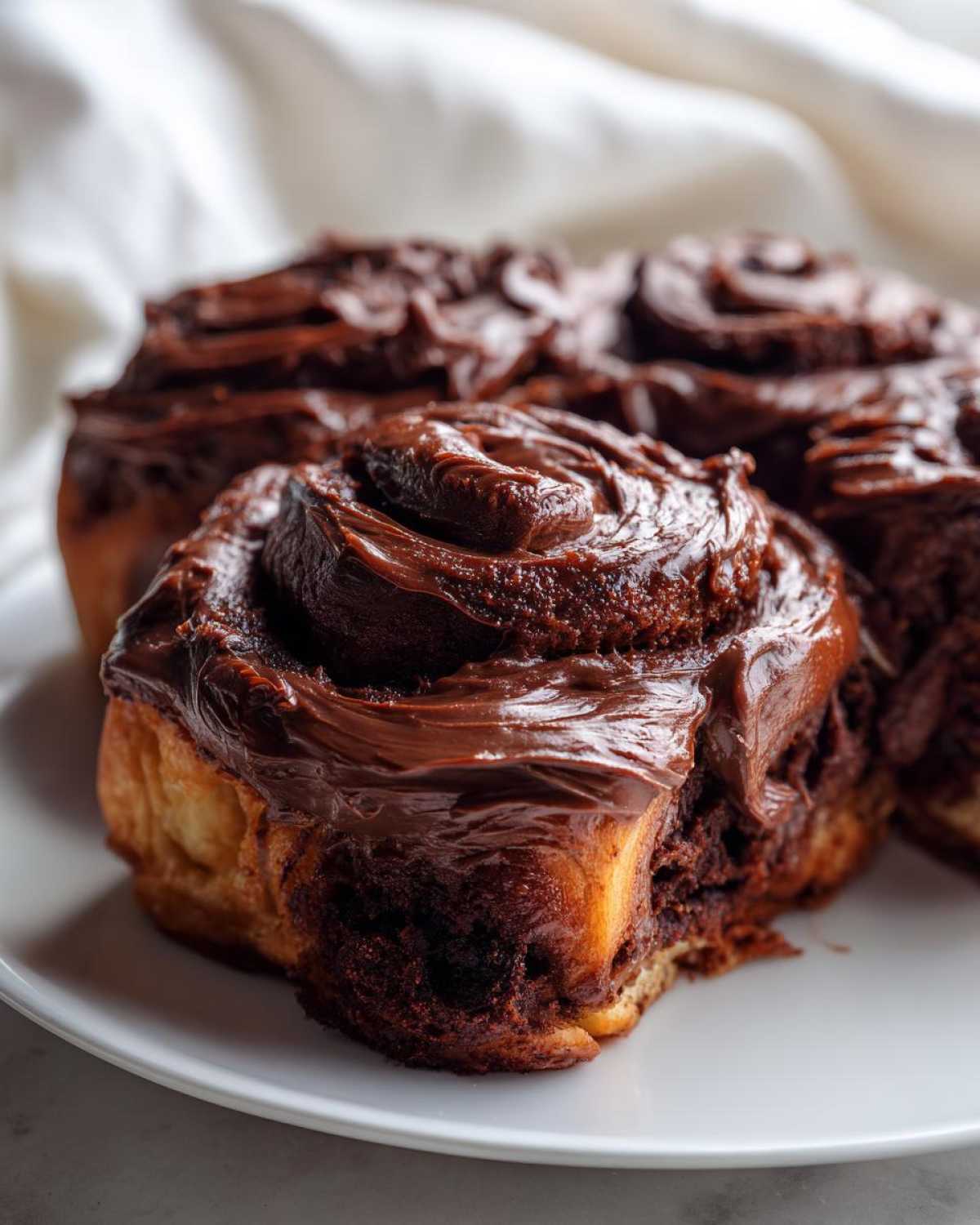 Close-up of a gooey chocolate cinnamon roll topped with thick, glossy chocolate frosting on a white plate.