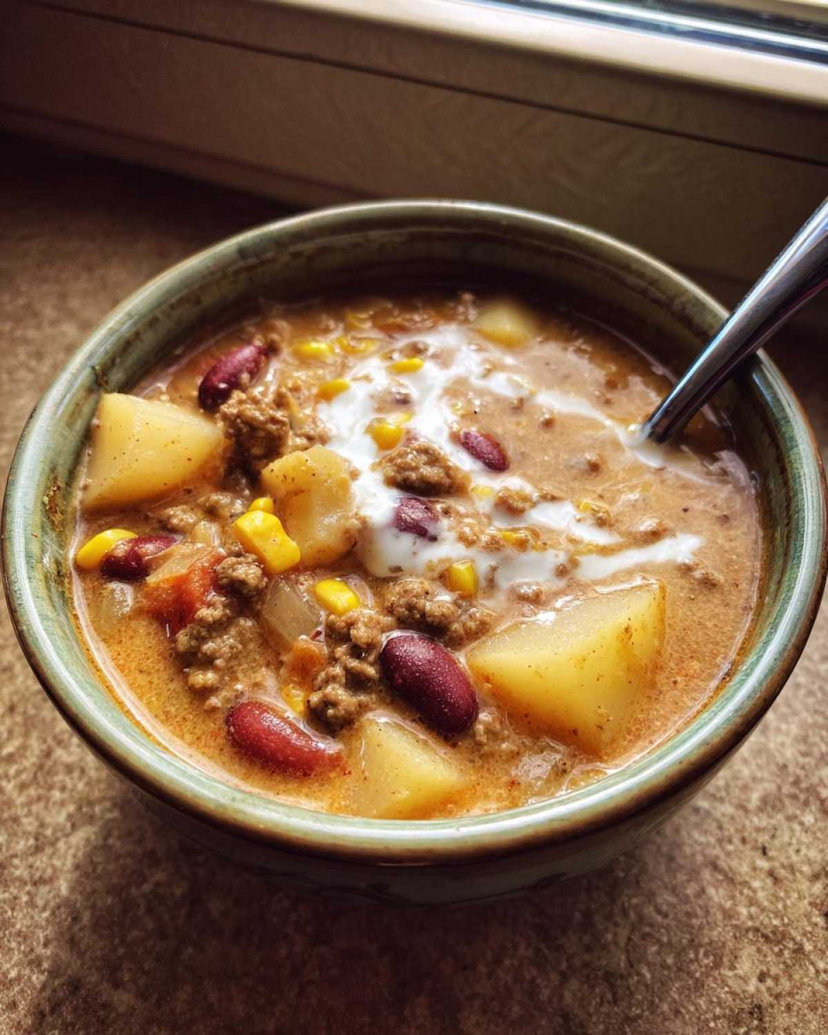Close-up of a hearty bowl of cowboy soup featuring ground meat, potatoes, corn, kidney beans, and a swirl of cream.