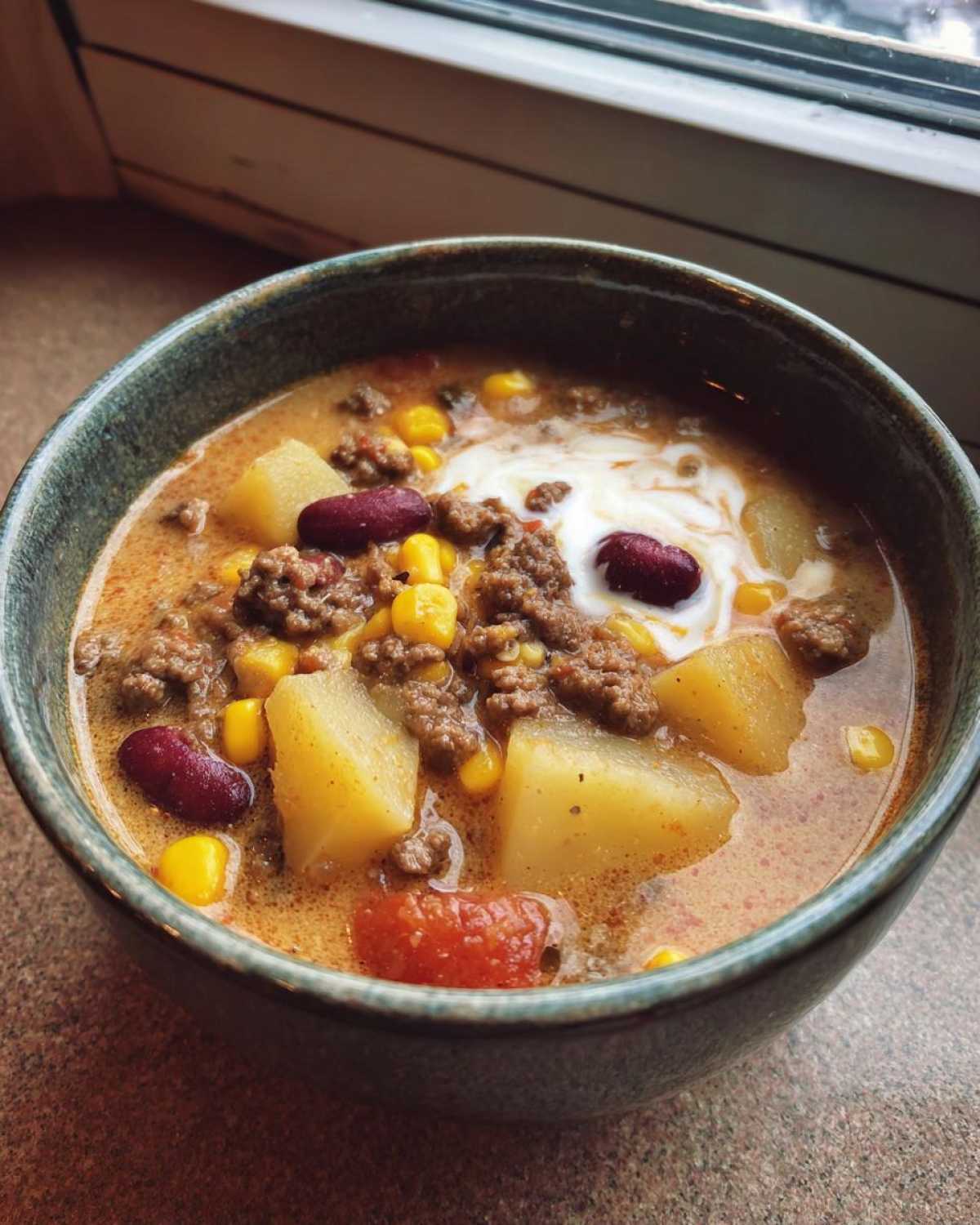 Close-up of a hearty bowl of cowboy soup featuring ground beef, potatoes, corn, and kidney beans, topped with sour cream.