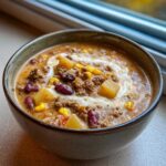 Close-up of a hearty bowl of cowboy soup topped with sour cream, showing ground beef, potatoes, corn, and kidney beans.