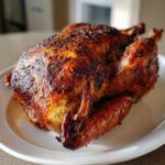 A whole roasted turkey with deeply browned, crispy skin seasoned with herbs, resting on a white plate.