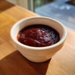 A small white bowl filled with thick, glossy homemade bbq sauce sitting on a wooden surface.