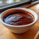 Close-up of thick, dark red homemade bbq sauce served in a small white ramekin on a wooden surface.
