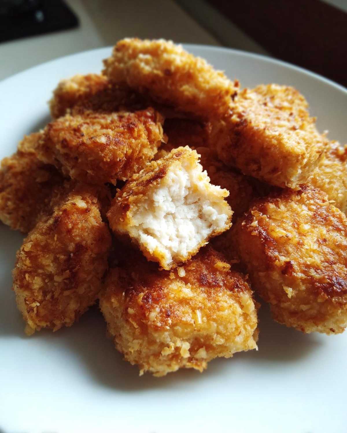 A pile of golden brown homemade chicken nuggets on a white plate, one nugget is broken open showing the white chicken meat inside.