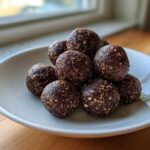 A close-up stack of dark brown, textured protein balls coated in seeds, resting on a white plate near a window.