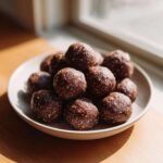 A close-up of a small bowl filled with homemade, dark brown protein balls sprinkled with seeds.
