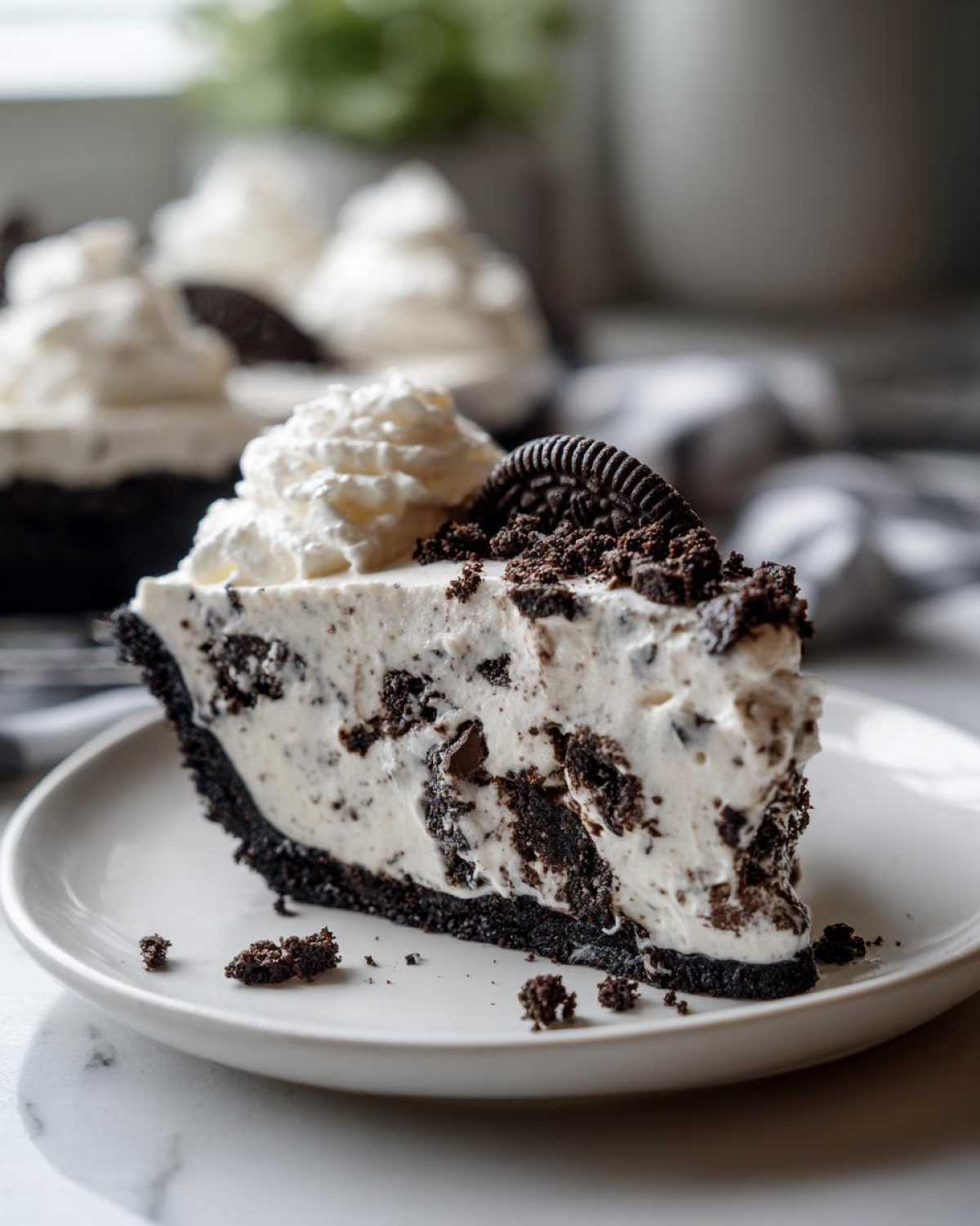 Close-up of a decadent slice of ice cream pie with a dark cookie crust, creamy filling mixed with cookie pieces, topped with whipped cream and an Oreo.